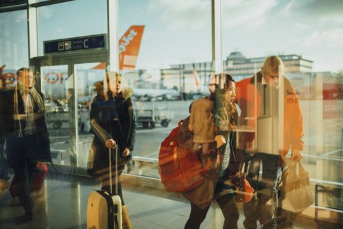 Passengers stroll through an airport terminal, pulling luggage behind them. Outside, planes are visible on the tarmac—a scene bustling with life yet mindful of safety to prevent any airport accidents. The bright reflections shimmer across the large glass windows.