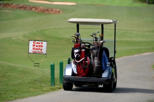 A golf cart filled with golf bags is parked on a path at a scenic golf course. A sign nearby reads "Cart Path Only This Hole," ensuring the carts, to avoid accidents, remain on designated paths. The lush green course stretches out with sand traps visible in the distance.