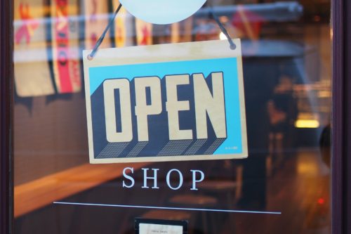 A brightly colored sign with the word "OPEN" hangs on a glass door, welcoming customers into the shop. The warm lighting and wooden furnishings give off a cozy vibe, reminiscent of an inviting corner in a bustling department store.