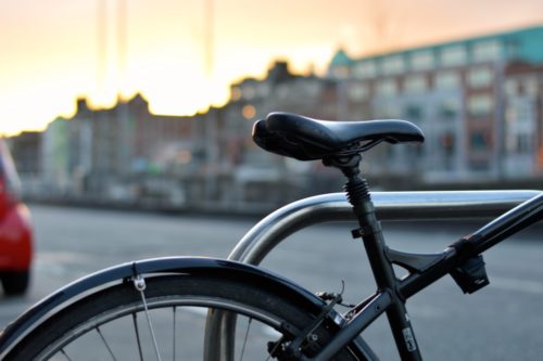 Close-up of a parked bicycle secured to a metal rack, with an urban street and buildings blurred in the background, creating a serene contrast to the chaos of a recent bicycle accident nearby. The sky is lit with a warm glow, suggesting either sunrise or sunset.