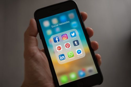 A hand holding a smartphone displays a folder labeled "Social Networks," featuring icons for Facebook, Instagram, Twitter, Google+, Pinterest, Tumblr, LinkedIn, WhatsApp, and Messenger. It's a colorful reminder of the challenge to avoid social media amid the vibrant digital landscape.