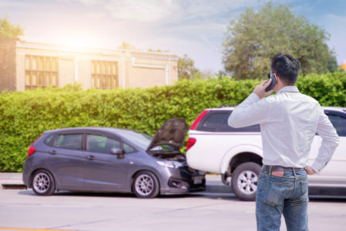 A man in a white shirt and jeans stands making a phone call near two cars involved in an accident. The gray car's hood is open, rear-ended by a white vehicle. With concern for possible injury, the scene unfolds against a backdrop of a lush green hedge and a building.