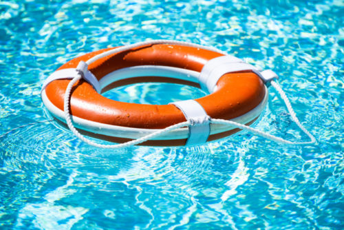 An orange life preserver with white straps floats on the clear blue water of a swimming pool, casting a shadow below. The sunlight creates rippling patterns on the pool's shimmering surface.
