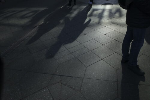Shadow of a person cast on a sunlit tiled pavement with geometric patterns. Partial view of a person wearing a dark coat on the right. Other shadows and faint outlines of a group of people are present on the ground.